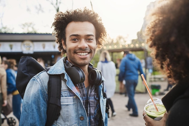 Hombre de piel oscura positivo y encantador con peinado afro caminando con su novia en el parque, sonriendo ampliamente a la cámara mientras está de buen humor. Hermanos que pasan tiempo juntos.