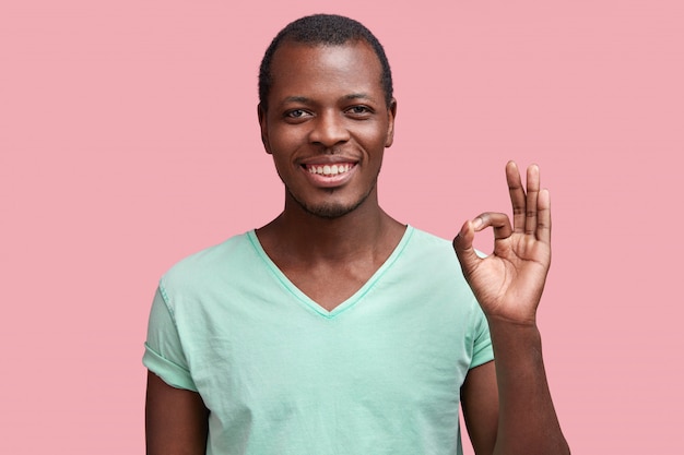 Hombre de piel oscura satisfecho con expresión feliz, gestos con la mano como muestra el signo de ok, demuestra que todo está bien, muestra aprobación, aislado sobre rosa