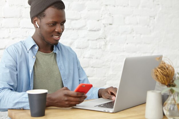 Hombre de piel oscura sonriente de aspecto moderno con sombrero usando auriculares inalámbricos mientras mira videos o series en línea en la computadora portátil, sentado en la mesa de café, enviando mensajes de texto a un teléfono móvil y bebiendo café