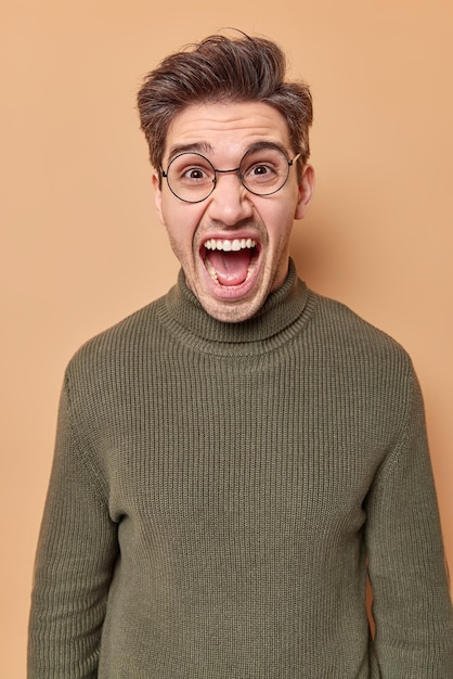 El hombre pierde los estribos, grita con la boca muy abierta, usa anteojos transparentes y un jersey casual aislado en beige. Concepto de personas y emociones