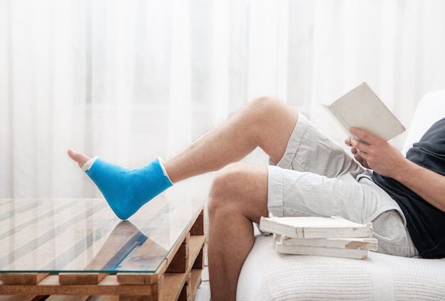 Un hombre con una pierna rota enyesada lee libros sobre un fondo claro del interior de la habitación.