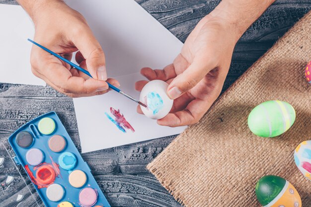 Hombre pintando un huevo con huevos de pascua en saco y pintura sobre fondo de madera oscura