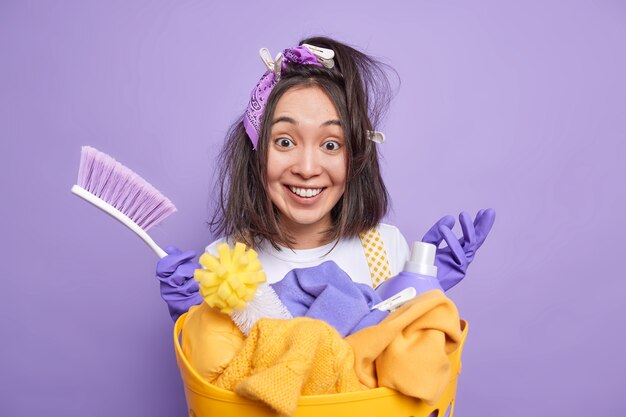 El hombre con pinzas para la ropa en el cabello sostiene el cepillo ocupado limpiando la casa sonríe positivamente se encuentra cerca de la canasta de lavandería usa detergentes aislados en violeta vivo