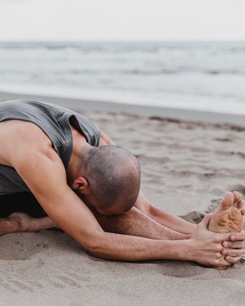 Foto gratuita hombre en la playa ejercitando posiciones de yoga en la arena