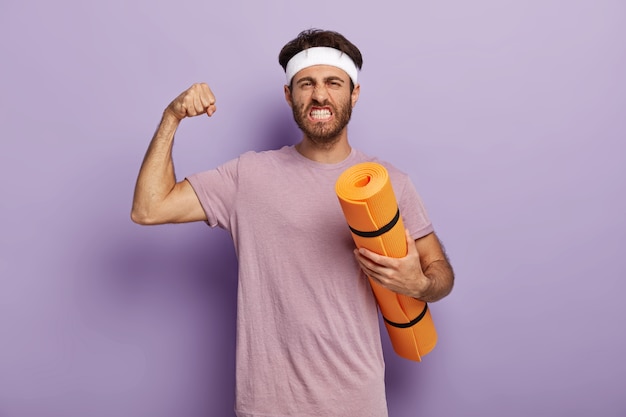 El hombre poderoso motivado se para con una colchoneta de fitness, disfruta del yoga como deporte y pasatiempo, levanta el brazo y muestra los músculos, aprieta los dientes, usa diadema, camiseta violeta. Equilibre su vida, lleve un estilo de vida saludable