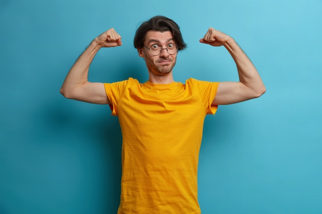 Hombre poderoso seguro de sí mismo levanta los brazos y muestra los músculos, demuestra el resultado de un entrenamiento regular, vestido con una camiseta amarilla y gafas, lleva un estilo de vida activo y saludable, siendo muy fuerte
