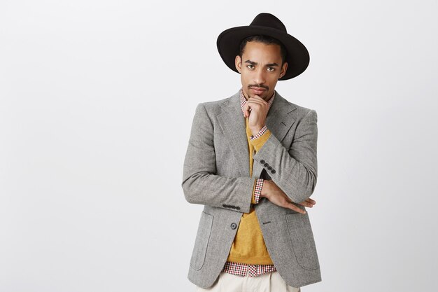 El hombre se pone serio cuando se trata de trabajo. Retrato de guapo joven enfocado de piel oscura con elegante sombrero y atuendo, sosteniendo la mano en la barbilla, mirando, tomando una decisión difícil sobre la pared gris