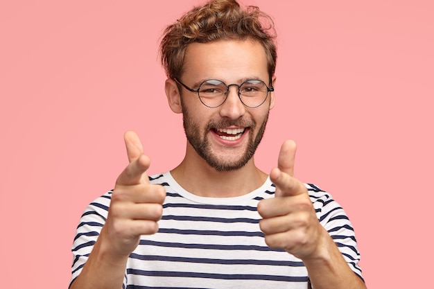 El hombre positivo sin afeitar le señala, tiene una expresión feliz, cabello rizado, cerdas, expresa su elección, usa ropa a rayas, aislado en la pared rosa. El hombre hace un gesto de pistola con los dedos. Tu eres mi tipo