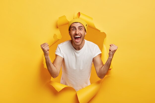 El hombre positivo aplaude con los puños cerrados, celebra la victoria, viste una camiseta blanca informal y un sombrero amarillo