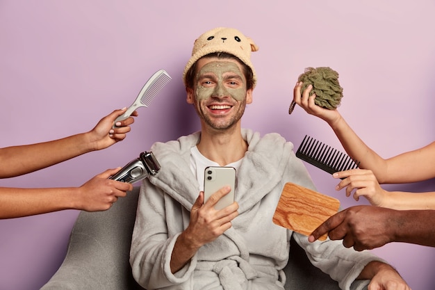 Hombre positivo bien arreglado usa albornoz, sombrero, usa teléfono inteligente después de tomar una ducha