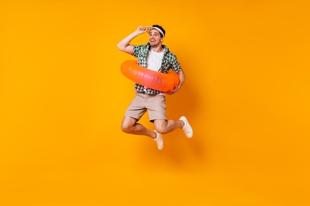 Hombre positivo está saltando sobre fondo naranja Retrato de chico con gorra y traje de verano con círculo inflable