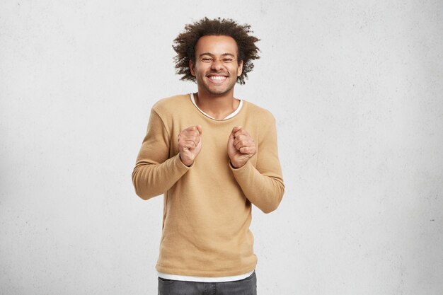 Hombre positivo feliz con pelo rizado tupido, levanta los puños y sonríe a la cámara, tiene una expresión encantadora