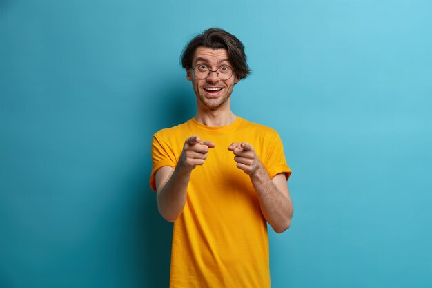 El hombre positivo feliz te señala con el dedo, elige a alguien, tiene una expresión alegre, dice bien hecho, elogia a un amigo por un buen trabajo o un resultado increíble, vestido de manera informal, aislado en una pared azul