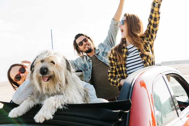 El hombre positivo y las mujeres sonrientes con las manos levantadas acercan al perro que se inclina hacia fuera del auto