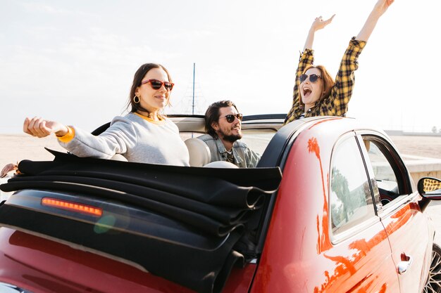 Hombre positivo que se sienta en el coche cerca de mujeres sonrientes con las manos levantadas que se inclinan hacia fuera del auto