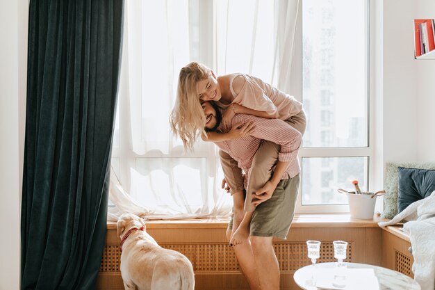 El hombre positivo en traje ligero lleva sobre su espalda a su amada novia. La pareja se divierte en su apartamento mientras su perro mira por la ventana.