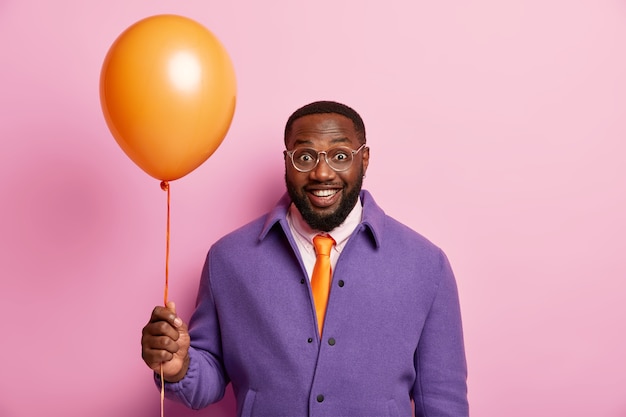 El hombre positivo viene a una reunión festiva con amigos, disfruta de las vacaciones, se para con un globo aerostático naranja, tiene buen humor