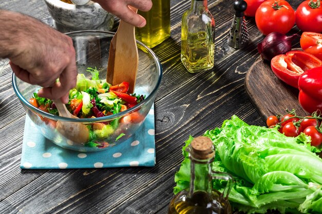 Hombre preparando ensalada con verduras frescas en una mesa de madera. Cocinar comida sabrosa y saludable. Sobre fondo negro. Comida vegetariana, saludable o concepto de cocina. De cerca