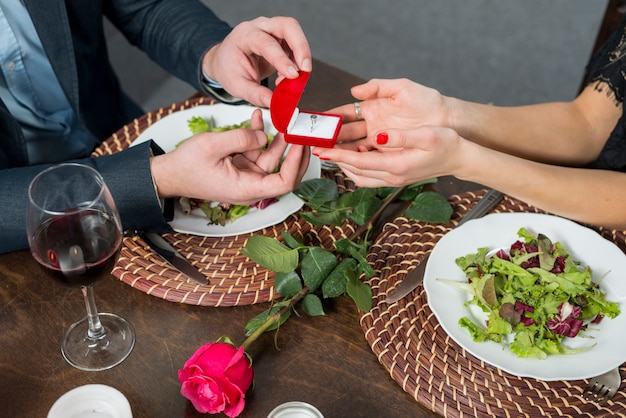 Hombre presentando caja de regalo a mujer en mesa con platos, rosa y vidrio