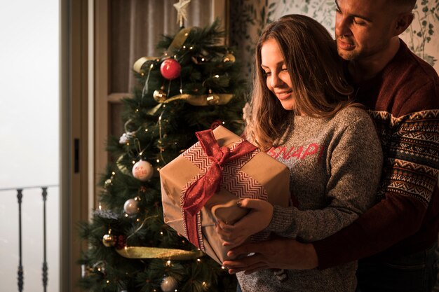 Hombre que abraza a la mujer alegre con la caja de regalo en suéteres cerca del árbol de navidad