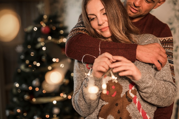 Hombre que abraza a la mujer alegre en suéteres cerca del árbol de navidad