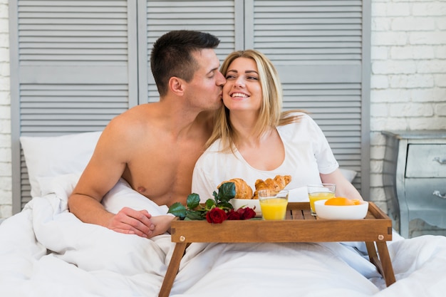 Hombre que besa a la mujer sonriente en cama cerca del desayuno a bordo