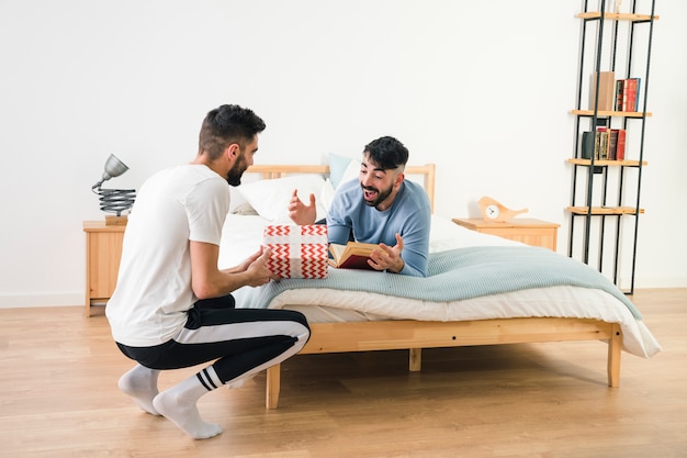 Hombre que da el regalo a su novio sorprendido acostado en la cama