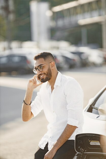 Hombre que habla con el móvil mientras espera el coche eléctrico. Estación de carga, coches ecológicos.