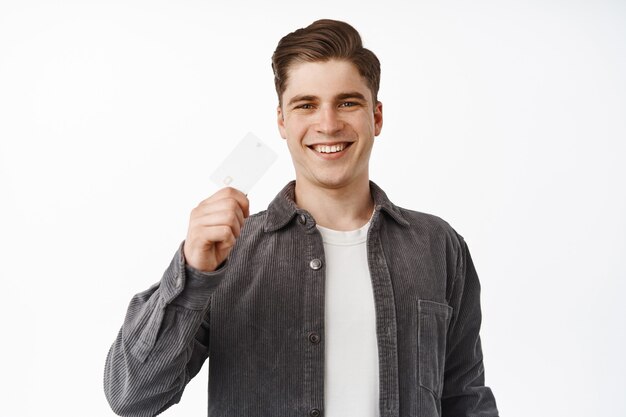 Hombre que muestra la tarjeta de crédito, paga sin contacto, se convierte en cliente del banco y se ve satisfecho, de pie con ropa informal en blanco