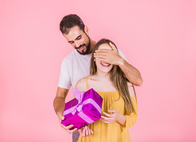 Hombre que oculta el ojo de la novia que le da el regalo sobre fondo rosado