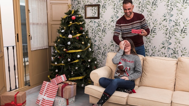 Hombre que presenta la caja de regalo y cierra los ojos a la mujer en el sofá cerca del árbol de Navidad