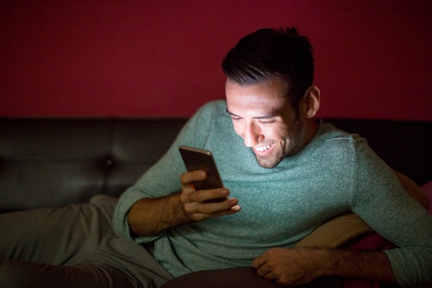 Hombre que ríe usando Smartphone en el sofá en la noche