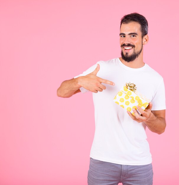 Hombre que señala el dedo en la caja de regalo con lazo dorado sobre fondo rosa