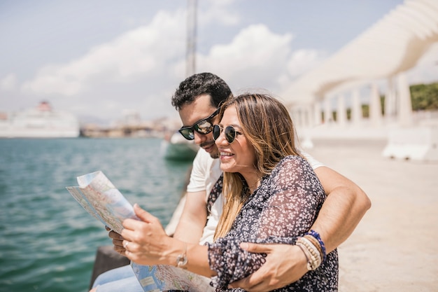 Hombre que se sienta en el embarcadero que abraza a su novia que busca la dirección en mapa