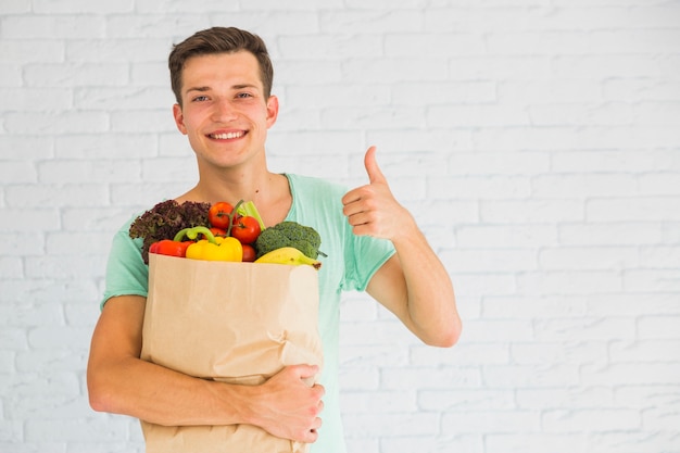 Hombre que sostiene la bolsa de papel marrón llena de frutas y verduras que muestra el pulgar en señal