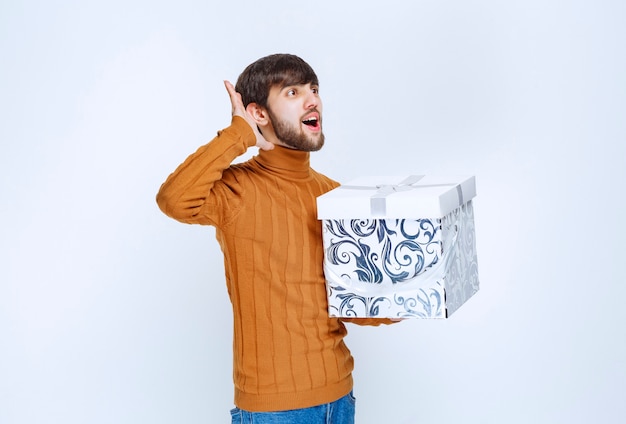 Hombre que sostiene una caja de regalo blanca con patrones azules que hacen un sonido para notar a alguien.