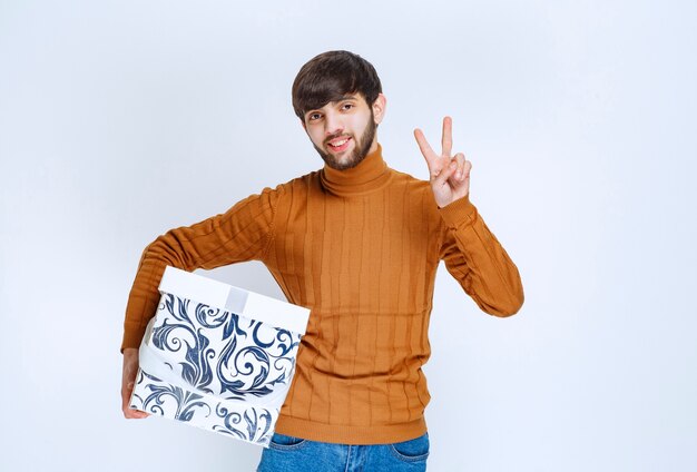 Hombre que sostiene una caja de regalo blanca con patrones azules y que muestra el signo de satisfacción.