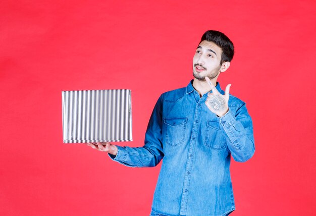 Hombre que sostiene una caja de regalo plateada en la pared roja y parece sorprendido y pensativo.