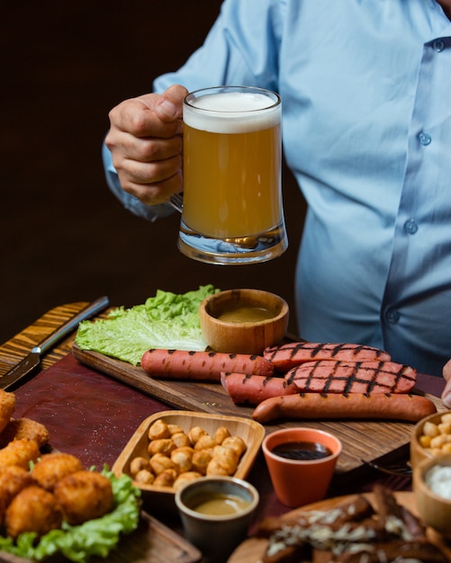 Foto gratuita hombre que sostiene la jarra de cerveza servida con salchichas a la parrilla, garbanzos, albóndigas crujientes