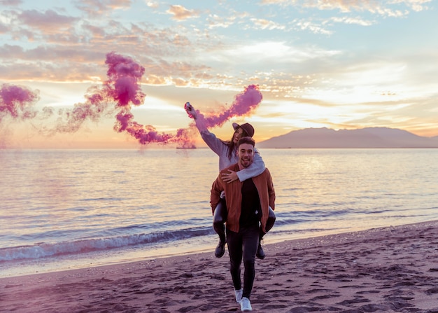 Foto gratuita hombre que sostiene a la mujer con la bomba de humo rosa en la espalda en la orilla del mar