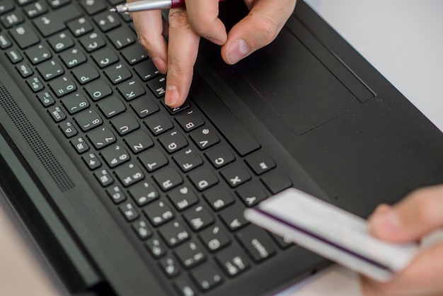 Hombre que sostiene la tarjeta de crédito en la mano y que incorpora el código de seguridad usando el teléfono elegante en el teclado de la computadora portátil, concepto en línea de las compras.