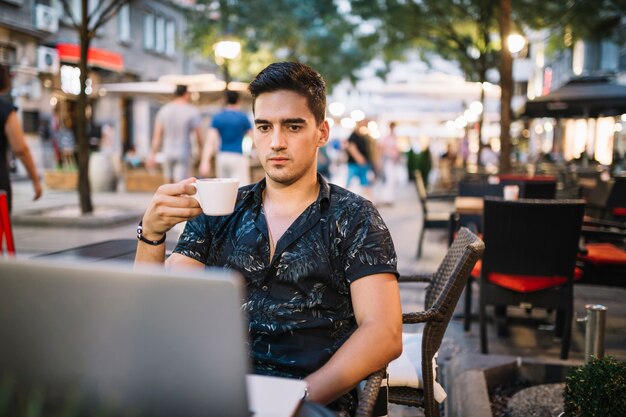 Hombre que sostiene la taza de café que mira la pantalla de la computadora portátil