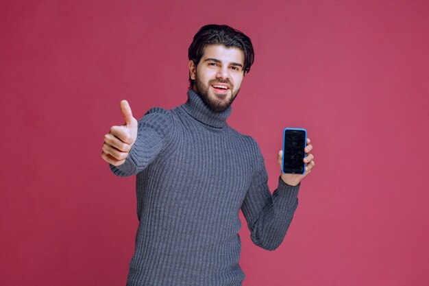 Hombre que sostiene un teléfono inteligente y hace un buen signo de mano.