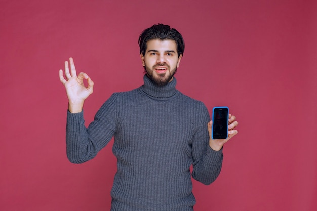 Hombre que sostiene un teléfono inteligente y hace un buen signo de mano.