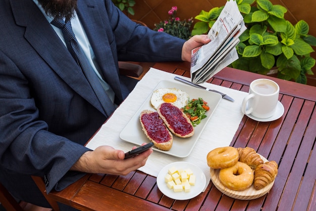 Hombre que sostiene el teléfono inteligente y el periódico durante el desayuno