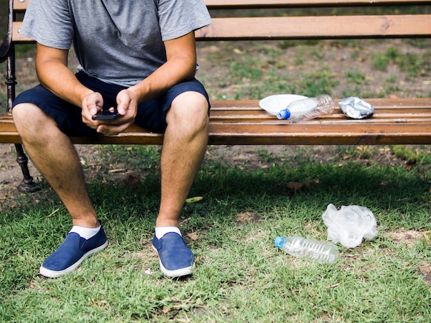 Hombre que usa el teléfono celular sentado en un banco cerca de la basura plástica en el parque