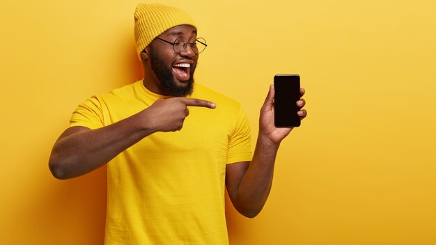 Hombre de raza negra con cerdas gruesas, apunta a un dispositivo de teléfono inteligente, muestra una pantalla en blanco para su contenido promocional, usa un sombrero y una camiseta amarilla informal, anuncia un nuevo dispositivo para los clientes