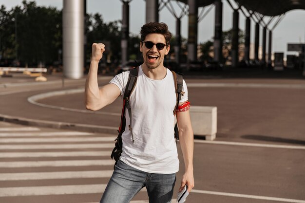 El hombre se regocija y sonríe. Guapo moreno con camiseta blanca y gafas de sol sostiene mochila y pasaporte cerca del aeropuerto.