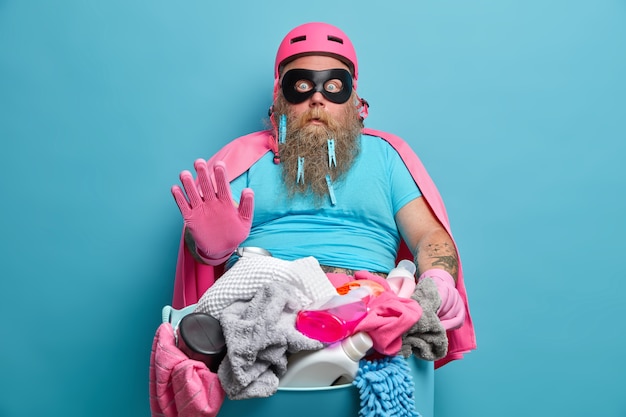 Hombre regordete aturdido con barba espesa hace gesto de parada prepara la ropa para la colada vestida con máscara de héroe y capa tontas alrededor