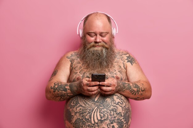 Hombre regordete complacido con cuerpo desnudo tatuado, gran barriga, escucha música en auriculares, tiene celular, descarga canciones en la lista de reproducción, aislada en la pared rosa. Personas, sobrepeso, concepto de hobby